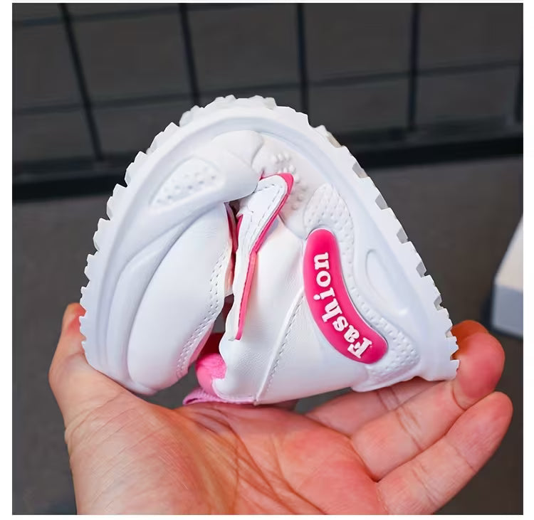 White kids sneaker with pink 'Fashion' tag, soft sole, held in hand for size view