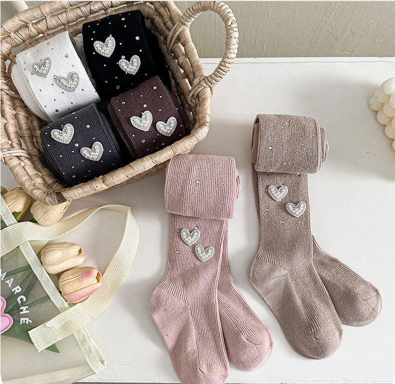 Kids' heart-embellished tights in pink, beige, black, and brown, displayed in a basket with flowers.