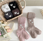 Kids' heart-embellished tights in pink, beige, black, and brown, displayed in a basket with flowers.
